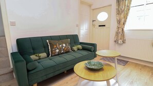 A green sofa with floral cushions, a small wooden table with a bowl, and a white radiator. - Crown Cottage (Belper)