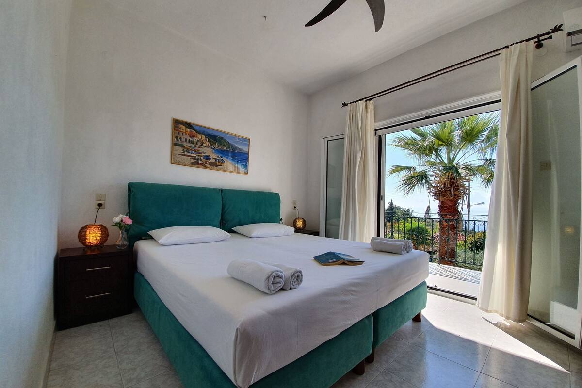 Una camera da letto con un letto, un comodino, un quadro e un balcone con una palma.