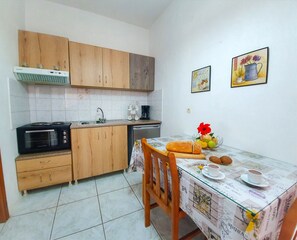 A compact kitchen with wooden cabinets, a built-in oven, and a dining table with a floral tablecloth. - Giovanni Apartments (Igoumenitsa)