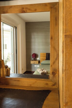Una stanza con pavimento in legno, una finestra, un letto con biancheria verde, un piccolo scaffale con una lampada e una testiera in legno.