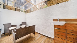 A patio with wicker furniture, a wooden cabinet, and a white brick wall. - West Kensington 1-Bed | Private Garden | Sleeps 4 (Greater London)