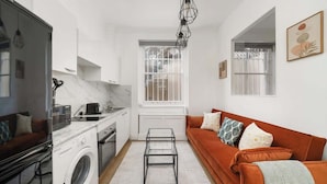 A compact living space with a kitchen, a washing machine, a sofa, and a dining area. - West Kensington 1-Bed | Private Garden | Sleeps 4 (Greater London)