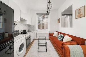 A compact living space with a kitchen, a washing machine, a sofa, and a dining area. - Stylish 1-Bed w/ Private Garden | West Kensington (Greater London)