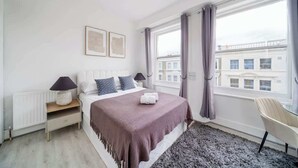 A bedroom with a bed, two bedside tables, a chair, a window with curtains, and a view of buildings. - West Kensington Studio | 3-Min Walk to Tube | Sleeps 2 (Greater London)