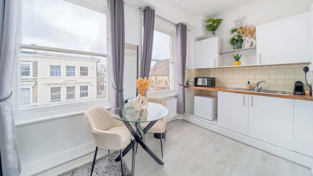 A modern kitchen with white cabinets, a glass dining table, and a view of buildings outside. - West Kensington Studio | 3-Min Walk to Tube | Sleeps 2 (Greater London)