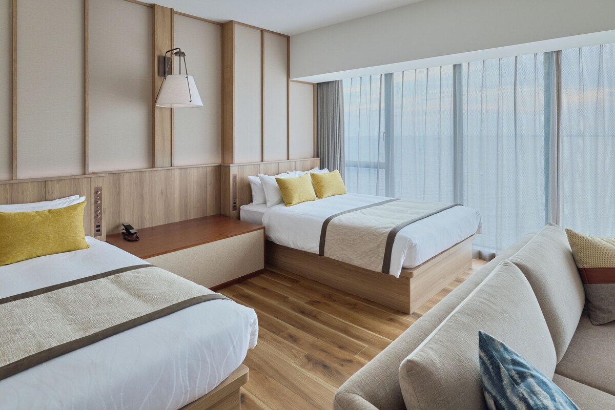 A hotel room with two beds, a sofa, a bedside table, and a window with curtains.
