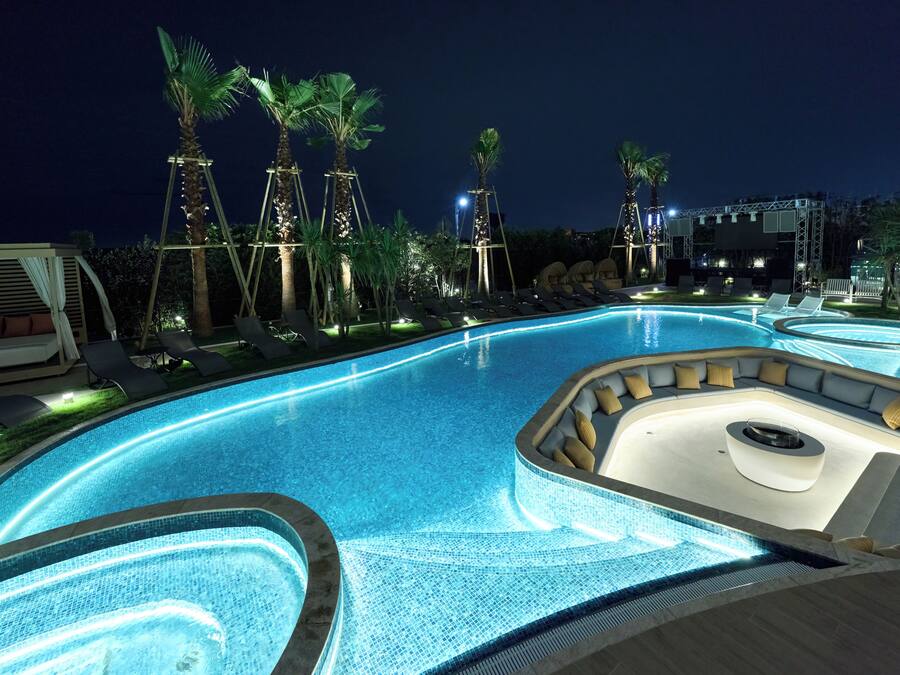Zona de piscina nocturna con sillones de descanso y palmeras.