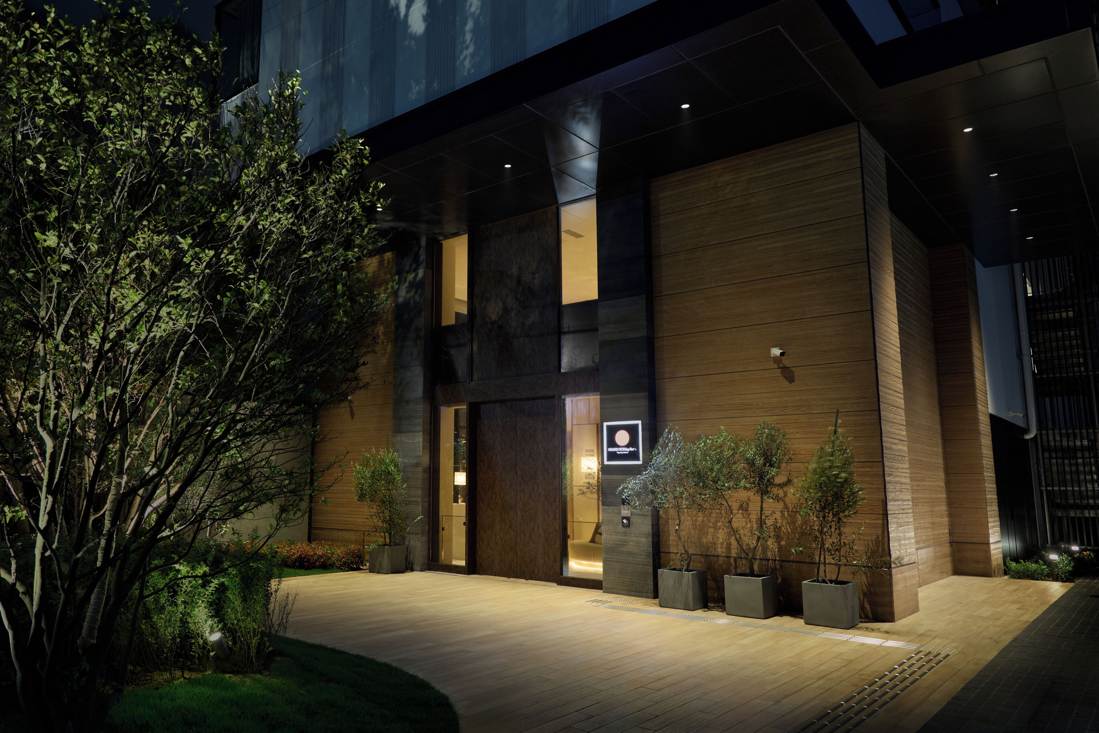 Un edificio moderno con una entrada amplia, plantas en macetas y un camino bien iluminado.