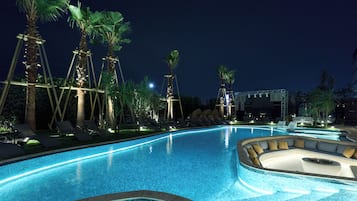 Una piscina nocturna con agua azul cristalina, rodeada de palmeras y sillas de descanso.