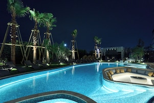 A nighttime swimming pool with clear blue water, surrounded by palm trees and lounge chairs.