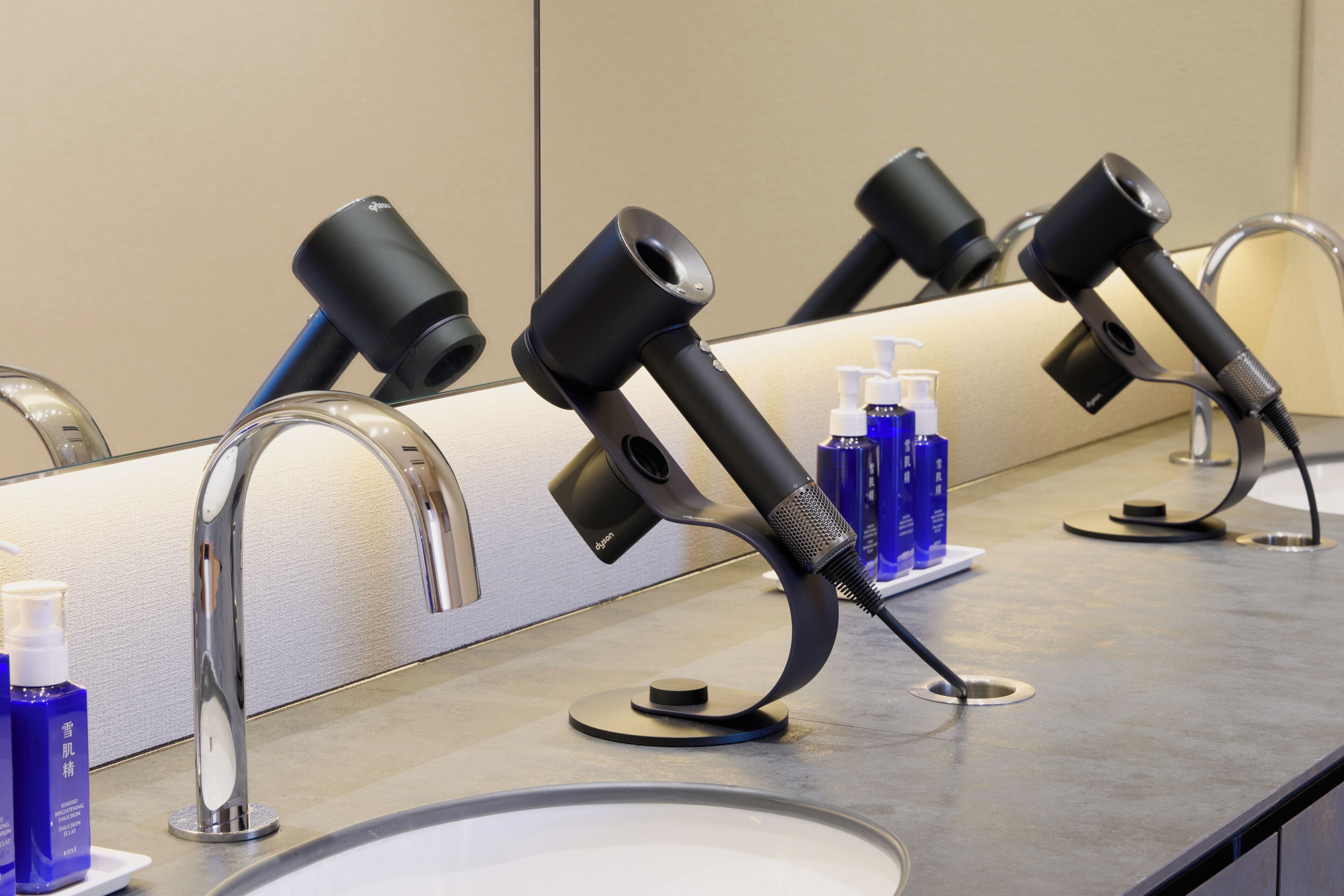 A bathroom with multiple hair dryers mounted on a wall, a sink, and several bottles of toiletries on the counter.