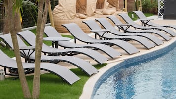 Zona junto a la piscina con sillones de descanso y una mesa pequeña, rodeada de césped y árboles.