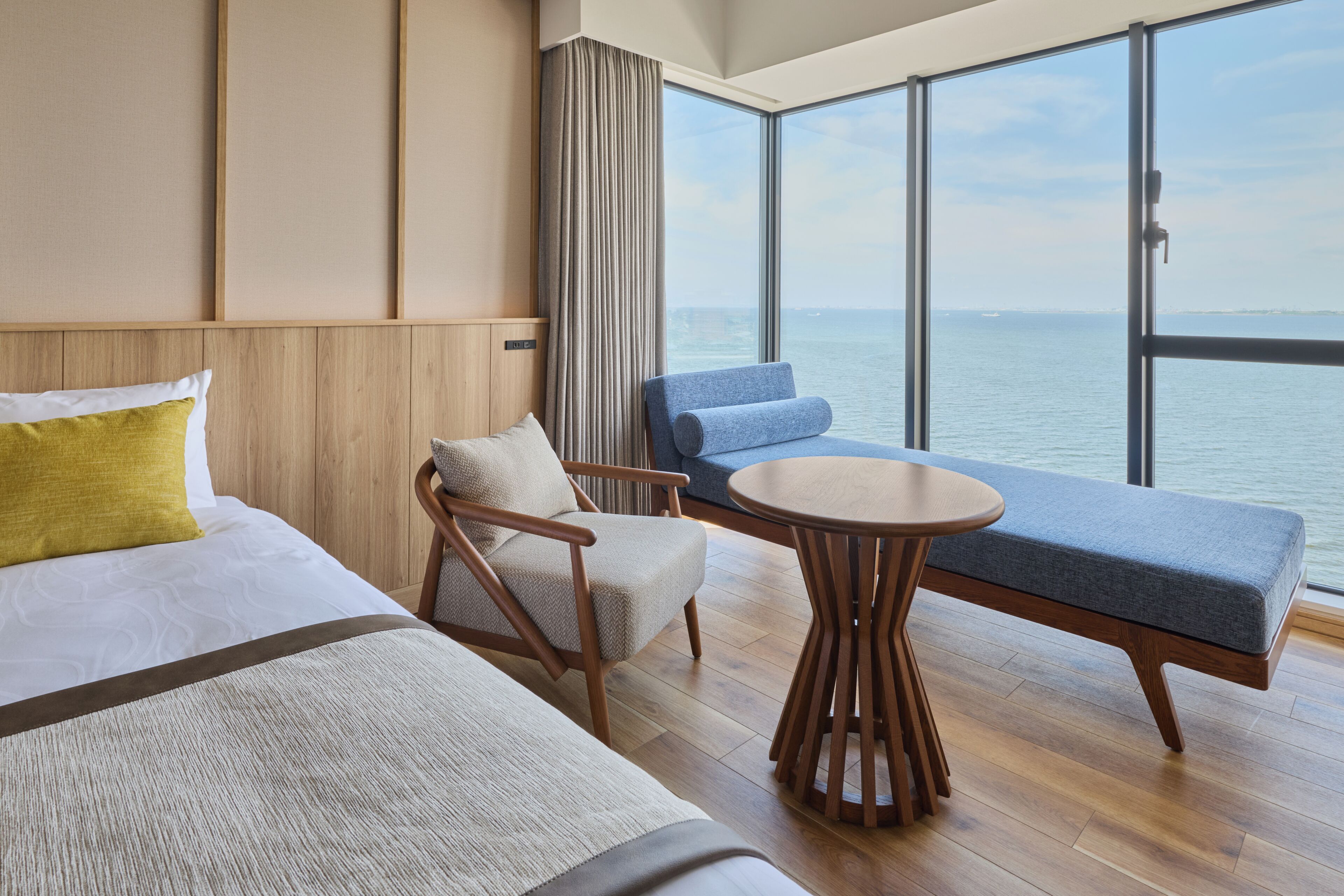 Quarto de hotel com uma janela grande, uma cama, uma poltrona, um sofá e uma mesa redonda pequena.