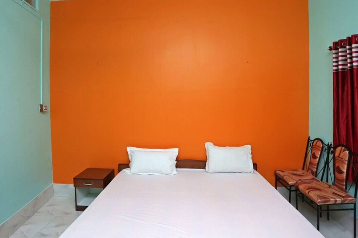 Quarto com parede laranja, cama com roupa de cama branca, mesa de cabeceira e duas cadeiras.