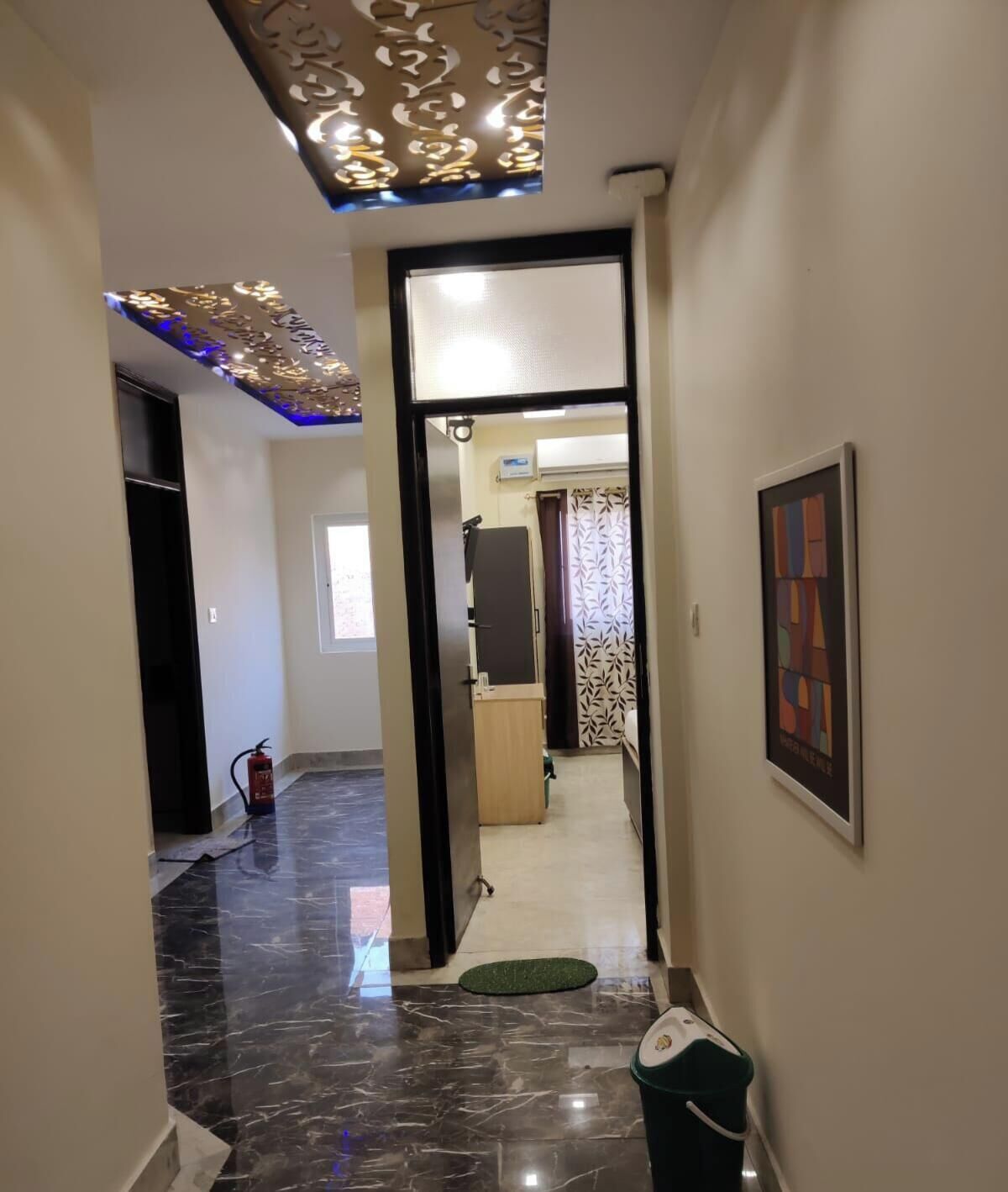 A hallway with a marble floor, a fire extinguisher, a green mat, a framed picture, and a ceiling light fixture.