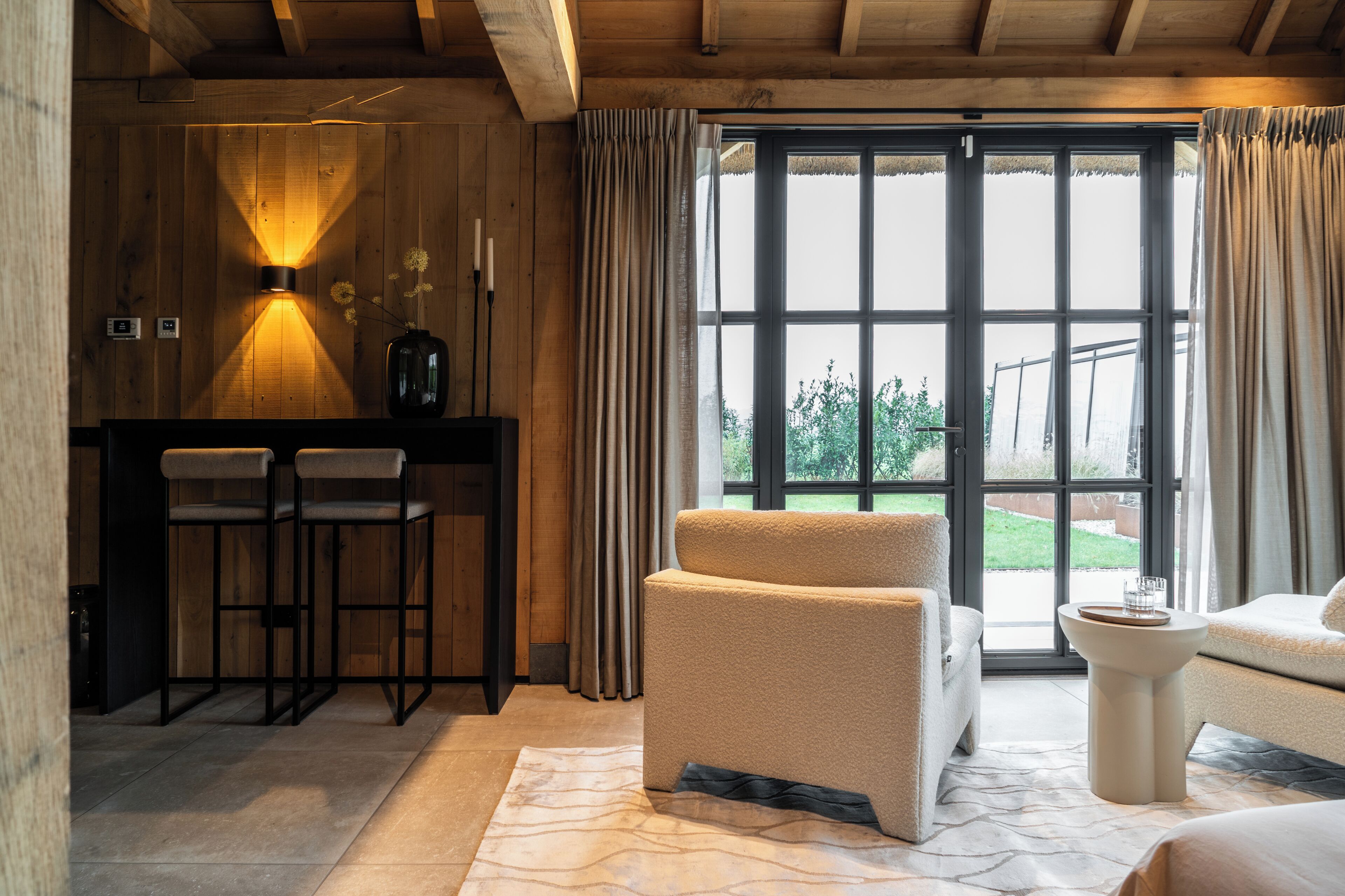 A room with a wooden interior, a bar area with stools, a sofa, and a large window with curtains.