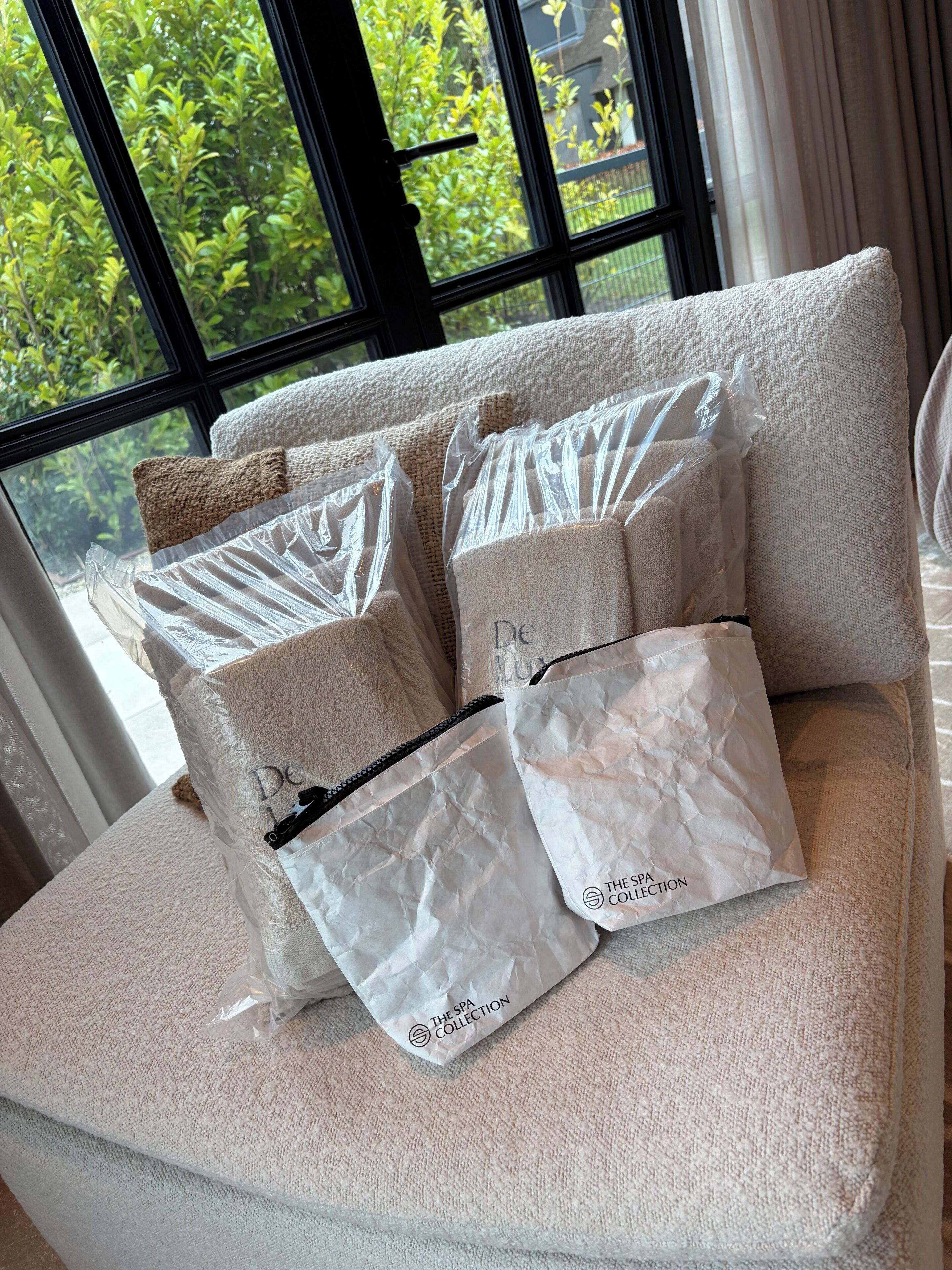 A sofa with multiple pillows and bags, some labeled 'The Spa Collection,' in a room with a large window and greenery outside.