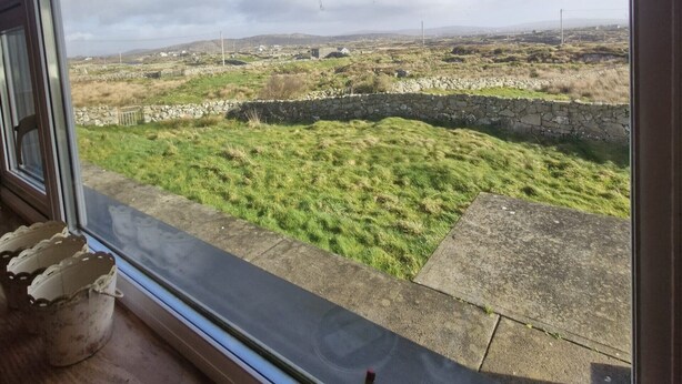 Vista desde el interior de una habitación con ventana que da a un paisaje con césped y muros de piedra.