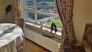 Una habitación con un ventanal grande, un radiador, una mesa con mantel de encaje y una silla.
