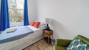 A bedroom with a bed, a nightstand, a lamp, a chair, and a window with curtains. - West Kensington 1-Bed | 3-Min Walk to Tube | Sleeps 4 (Greater London)