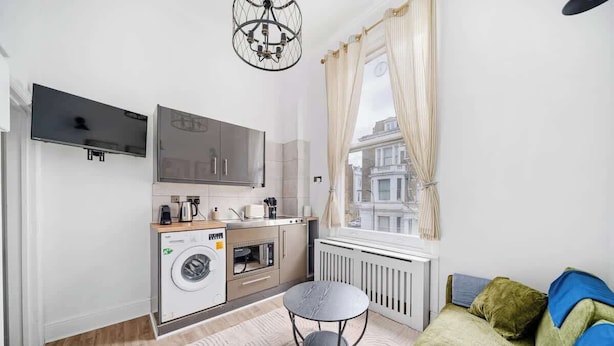 A compact living space with a kitchen, a washing machine, a dining table, and a sofa. - West Kensington 1-Bed | 3-Min Walk to Tube | Sleeps 4 (Greater London)