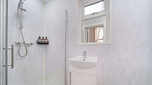 A bathroom with a shower, a sink, a mirror, and a shelf with bottles. - West Kensington 1-Bed | 3-Min Walk to Tube | Sleeps 4 (Greater London)