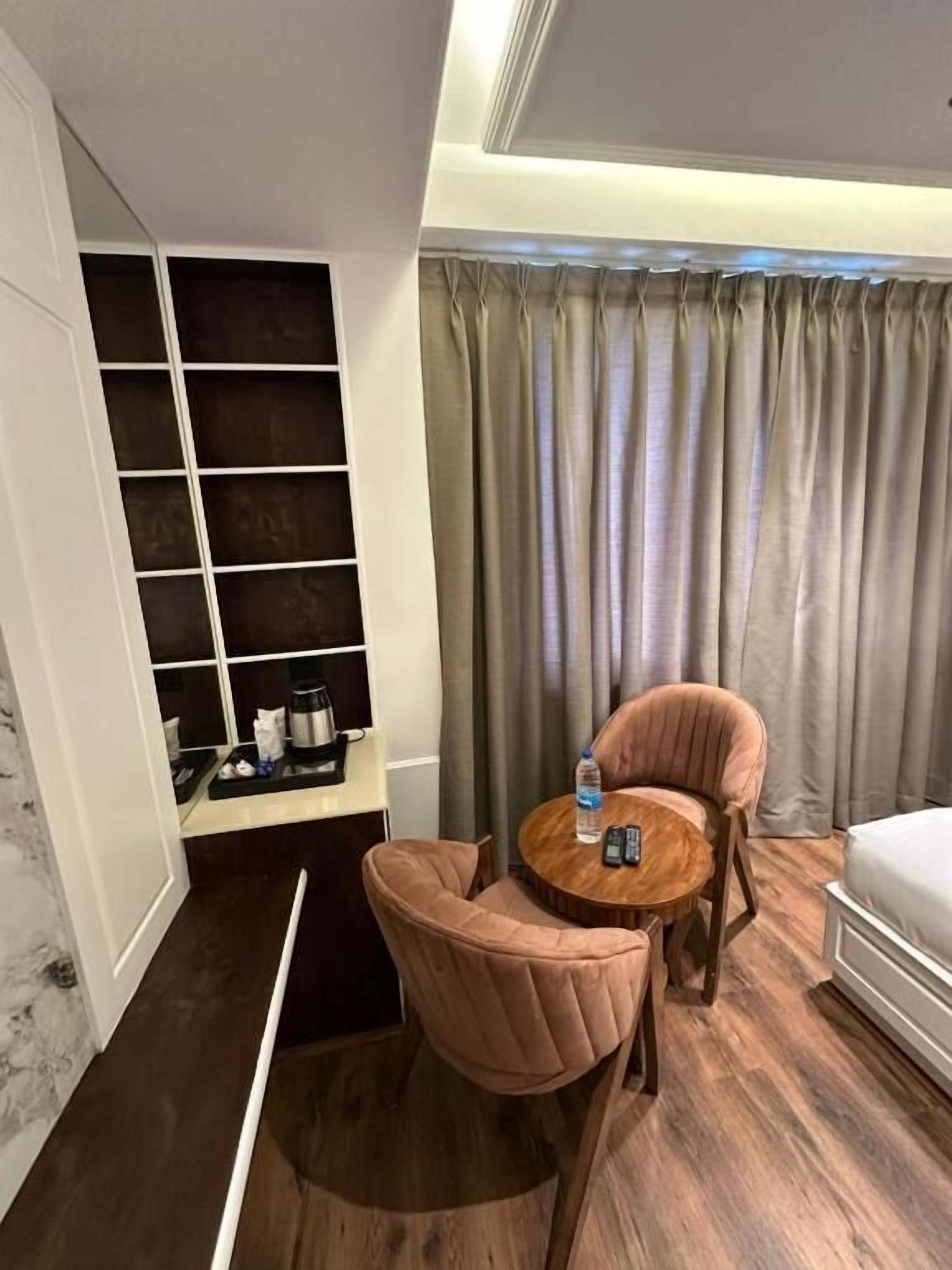 Habitación de hotel con piso de madera, una pequeña mesa redonda con una silla, un sofá, un minibar y un ventanal con cortinas.