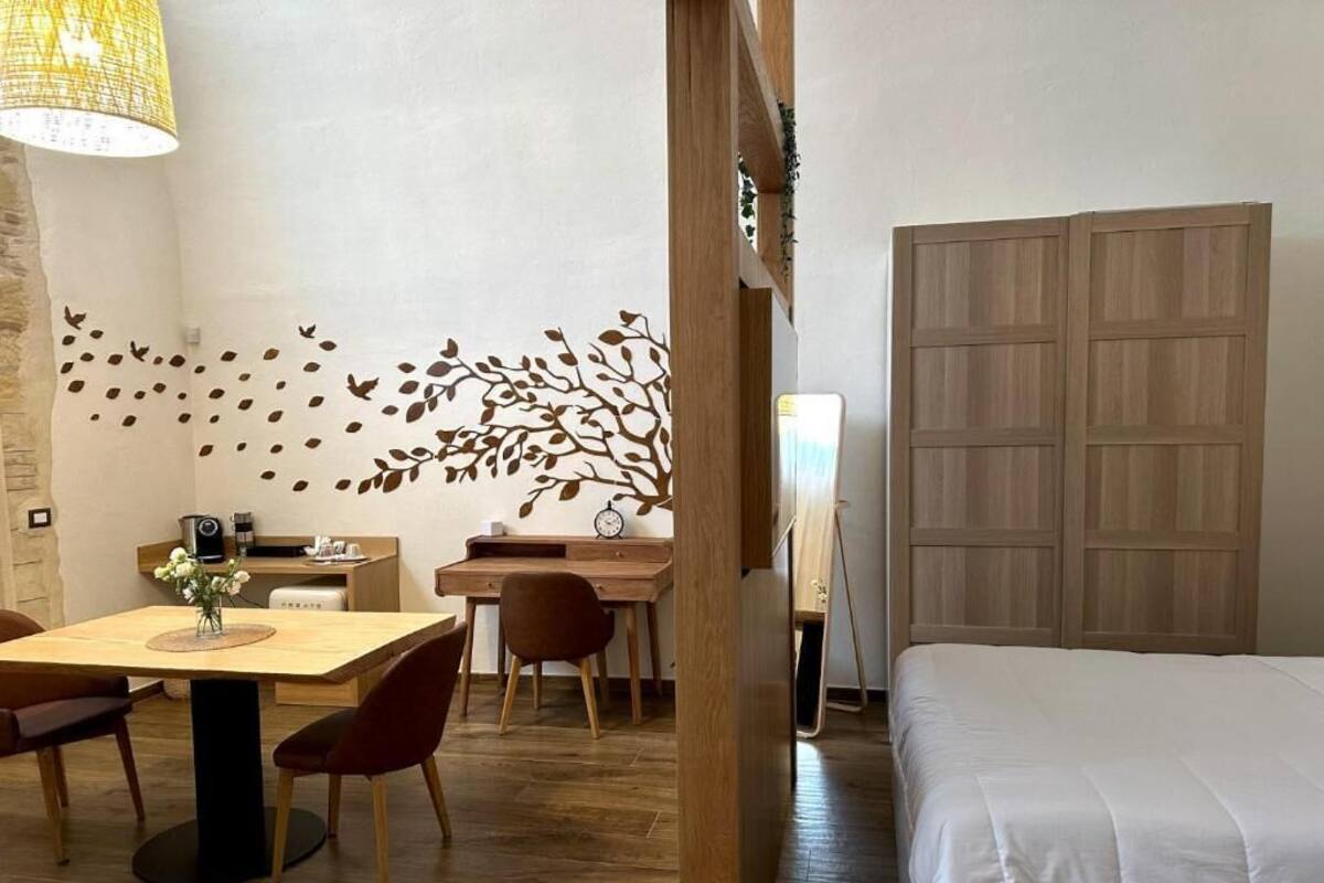 A hotel room with a bed, a desk with a chair, a dining table with chairs, a lamp, and a decorative wall with leaf patterns.