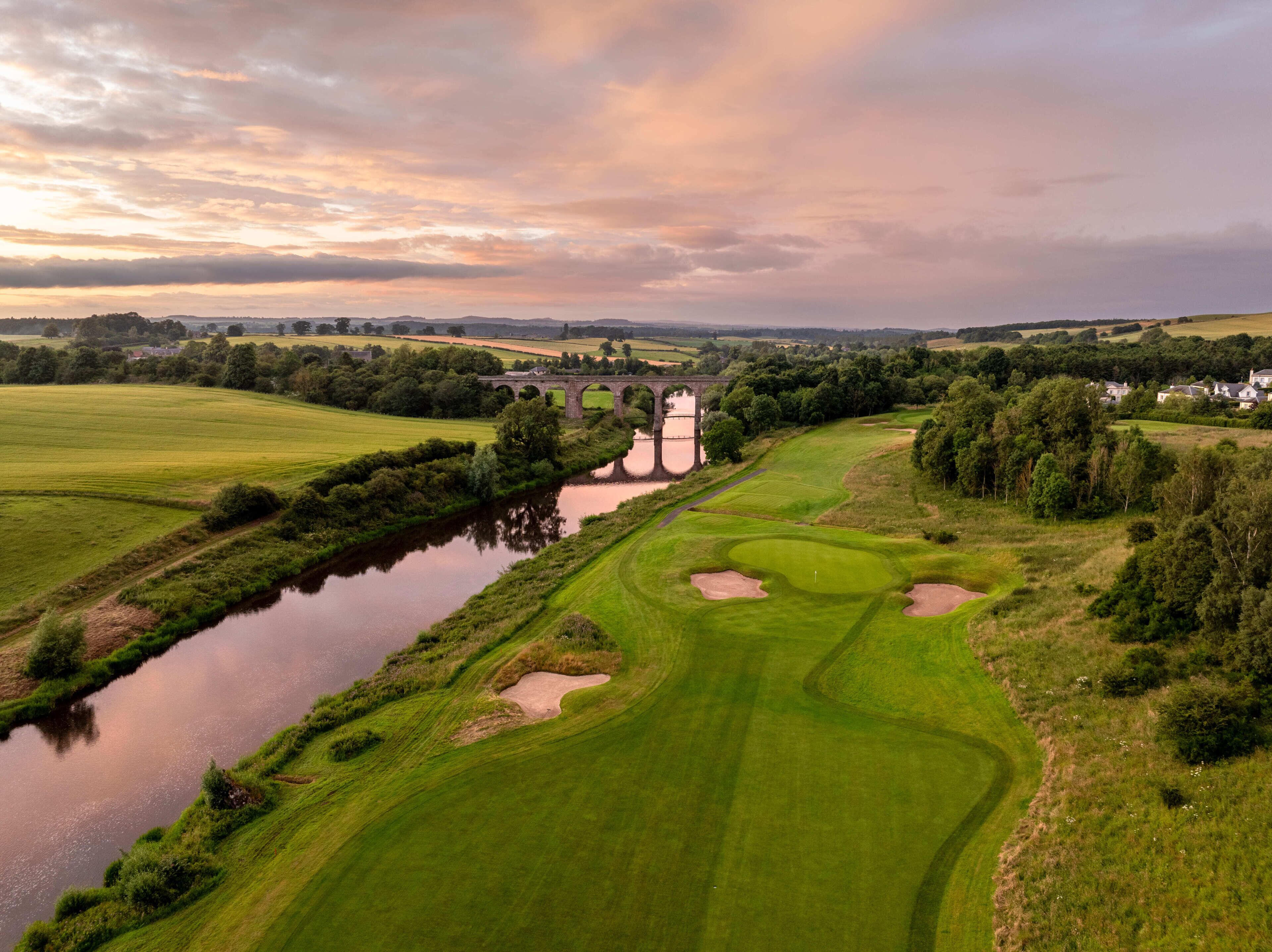 Un parcours de golf avec une rivière, un pont et des maisons en arrière-plan.
