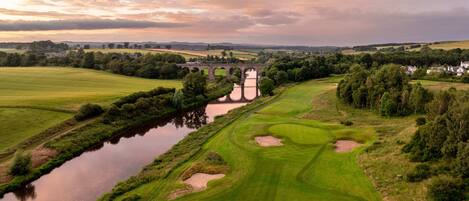 Un parcours de golf avec une rivière, un pont et des maisons en arrière-plan.