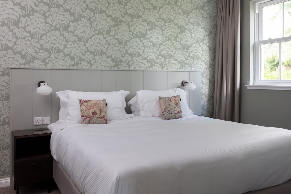 Una cama bien tendida con sábanas blancas y dos almohadones decorativos, una mesita de noche con lámpara, y una ventana con cortinas.