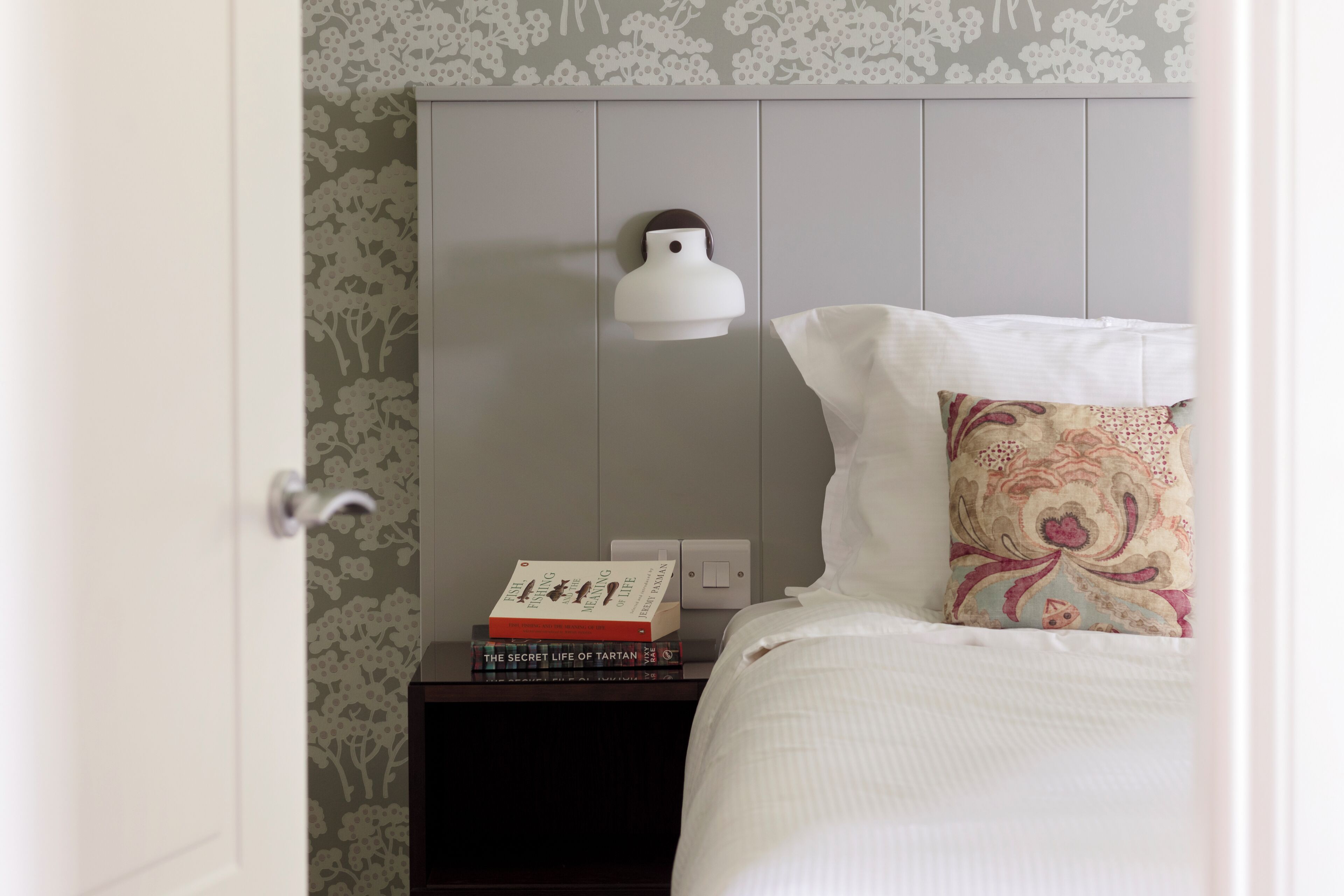 A neatly made bed with a patterned pillow, a bedside table with a lamp and books, and a door with a floral wallpaper design.