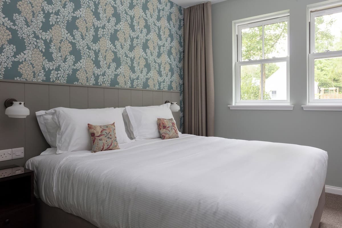 Una cama bien hecha con sábanas blancas y dos almohadones decorativos, una lámpara de pared y un ventanal con cortinas.