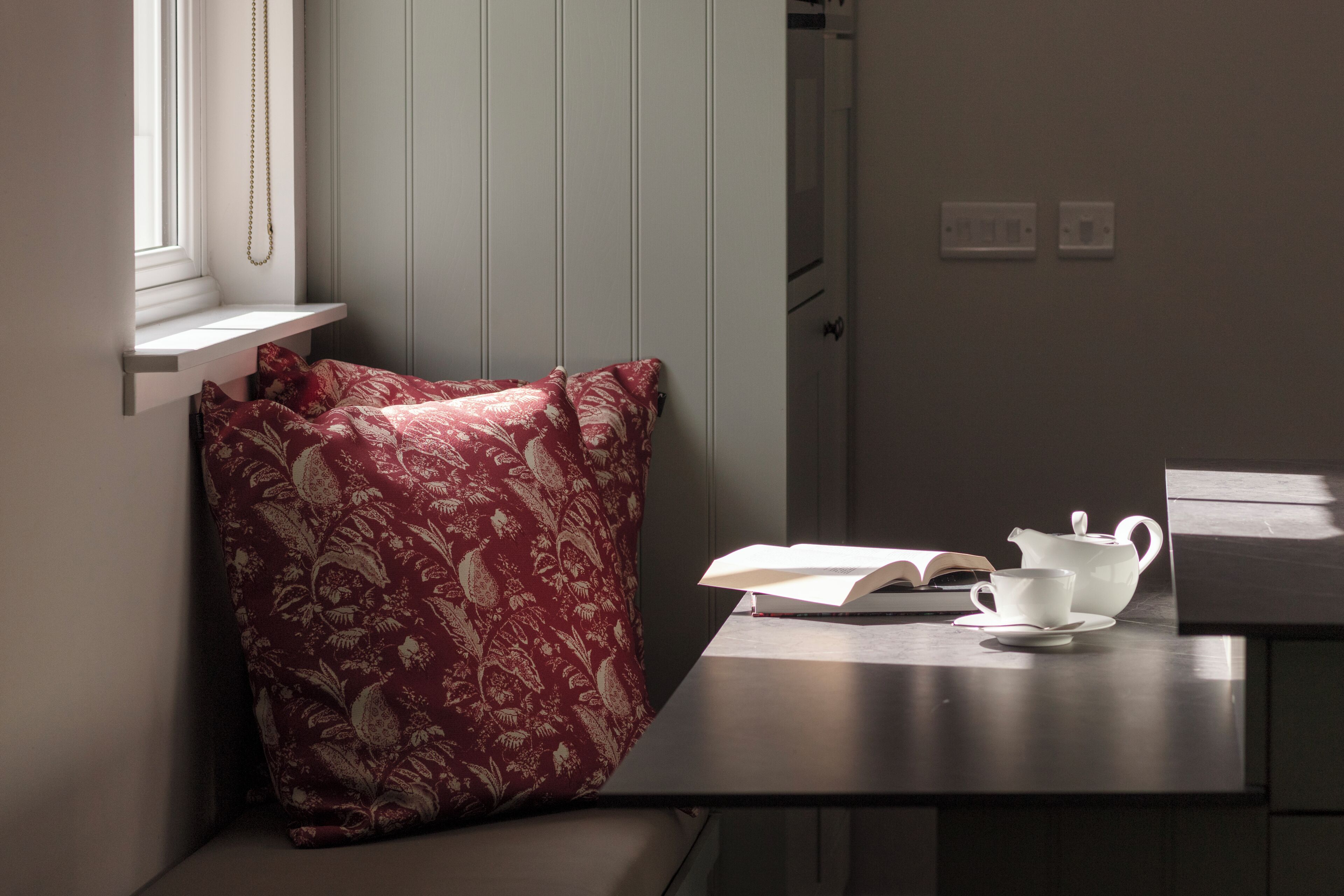 A corner room with a window, a cushion, a table with a book, a teapot, and a cup.