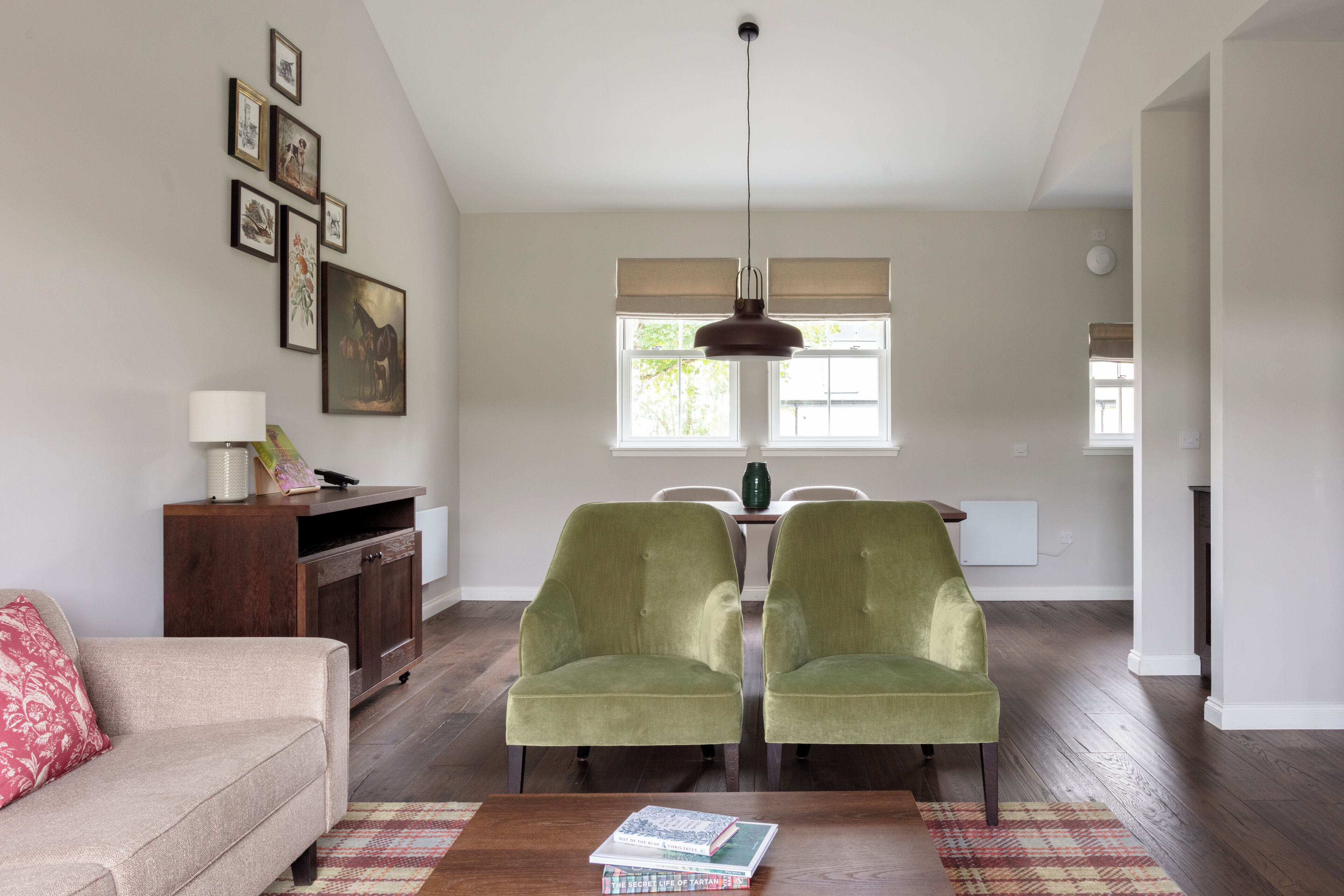 A living room with a sofa, two armchairs, a coffee table with books, a wooden cabinet, a hanging lamp, and a dining area with a table and chairs.