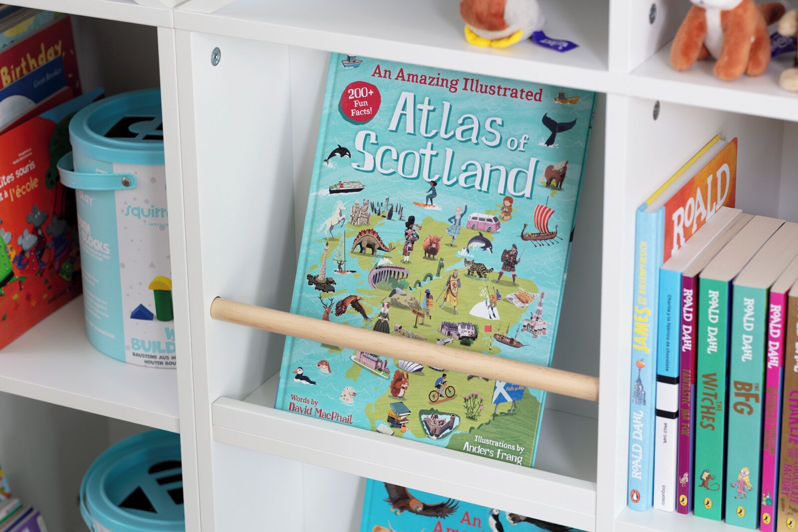 A bookshelf with a book titled 'Atlas of Scotland' by David MacPhail, surrounded by other books and colorful storage bins.