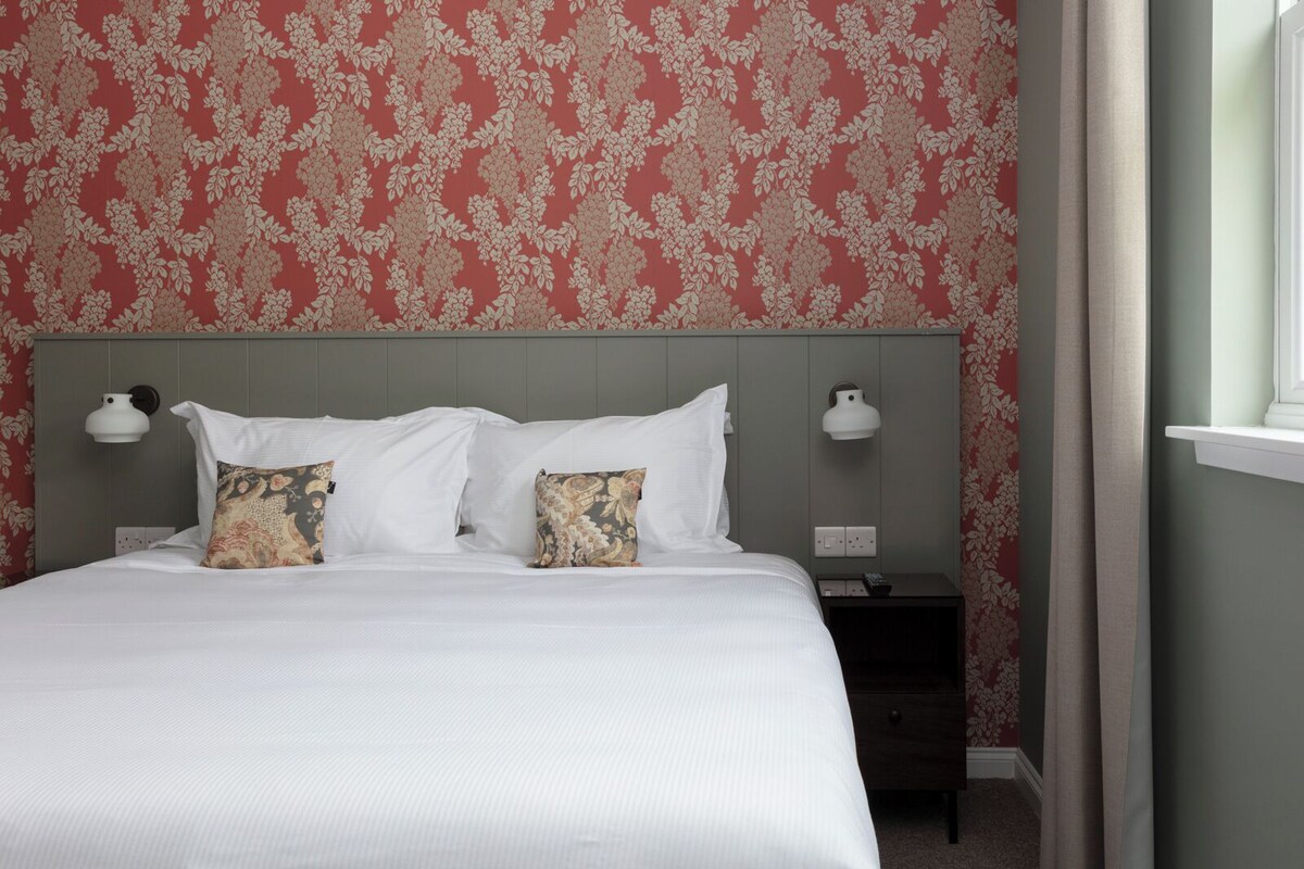 Una cama bien hecha con sábanas blancas y dos almohadas decorativas, una mesita de noche con lámpara y una pared con estampado floral.