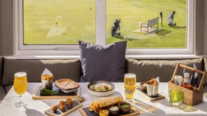 A dining table with food and drinks, a window with a view of a golf course, and a sofa with a pillow. - Roxburghe Cottages, Part of Destination by Hyatt (Kelso)