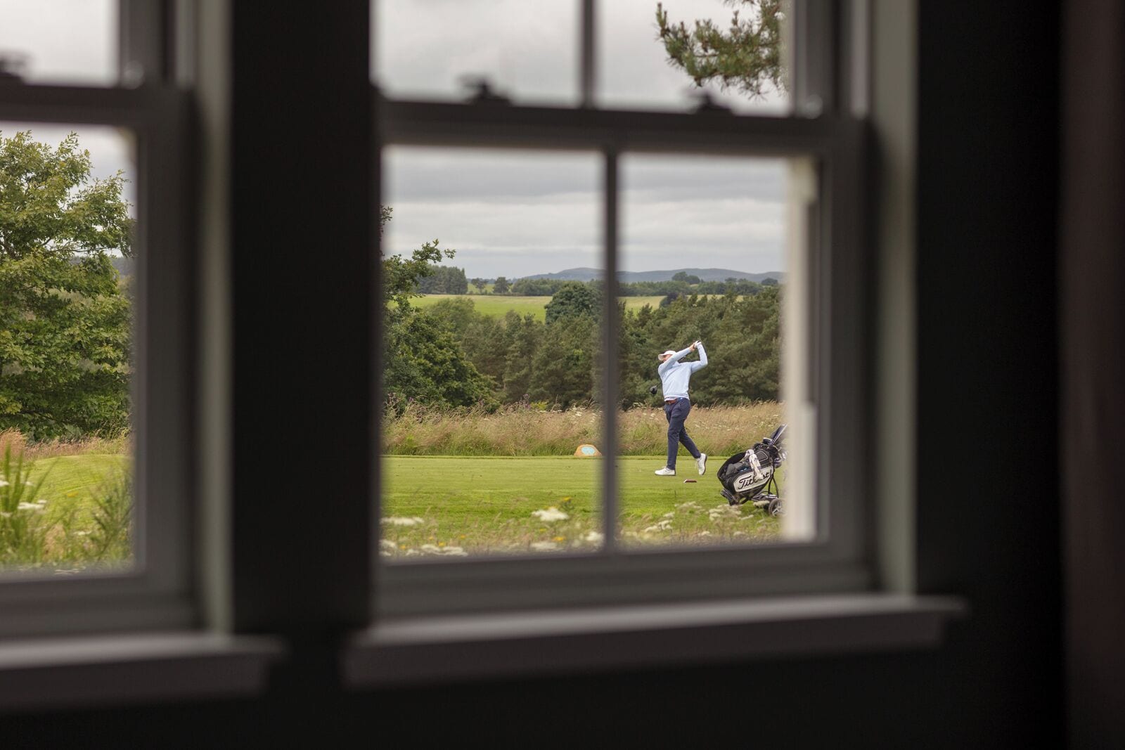 En vy från ett fönster ut över en golfbana, där en golfspelare svingar och en golfbagge ligger på marken.