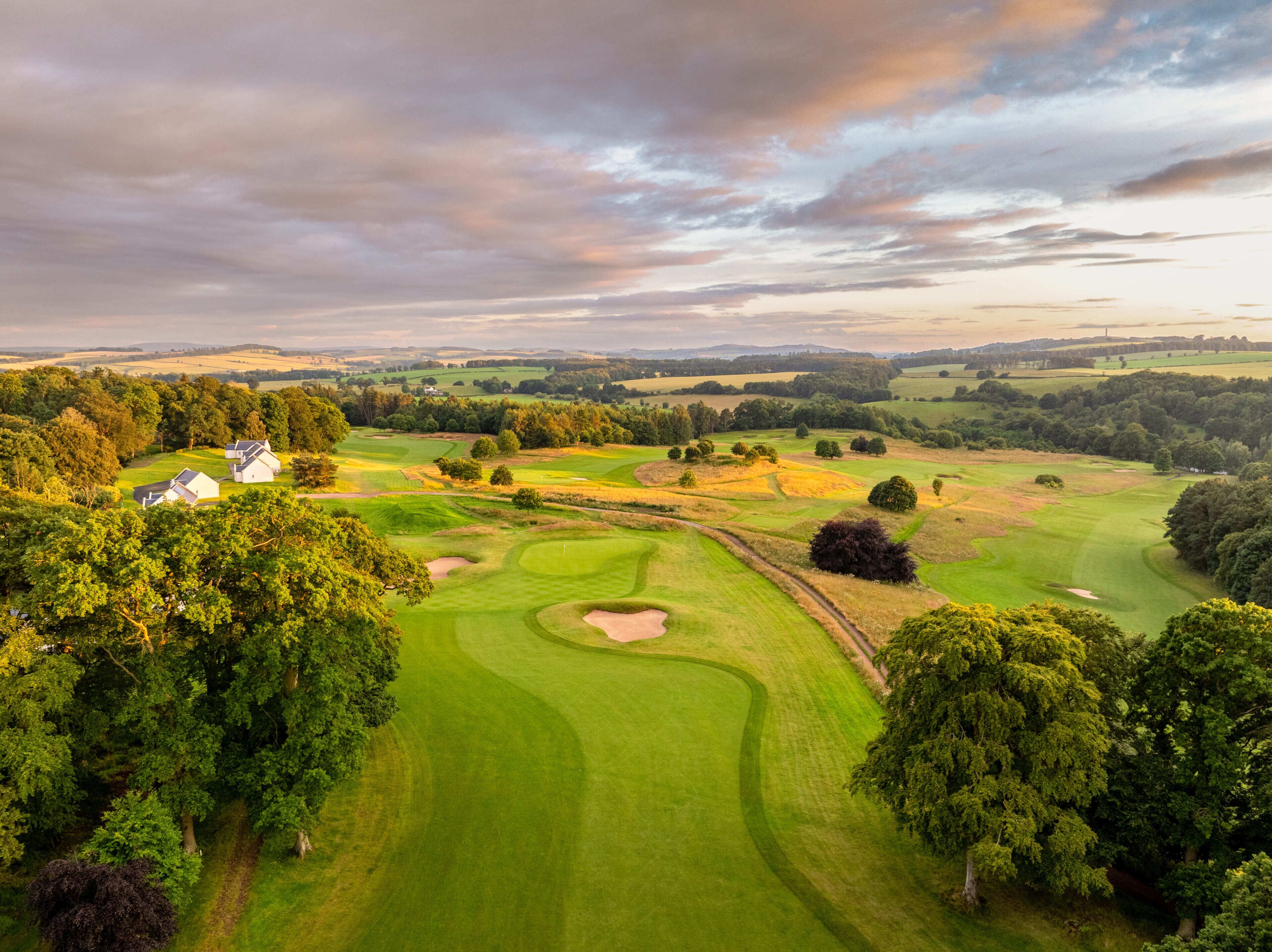 Un campo de golf con onduladas colinas verdes, árboles y algunos edificios blancos.