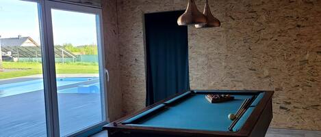 Une table de billard dans une pièce avec des poutres en bois, une grande fenêtre et une vue sur une piscine à l’extérieur.