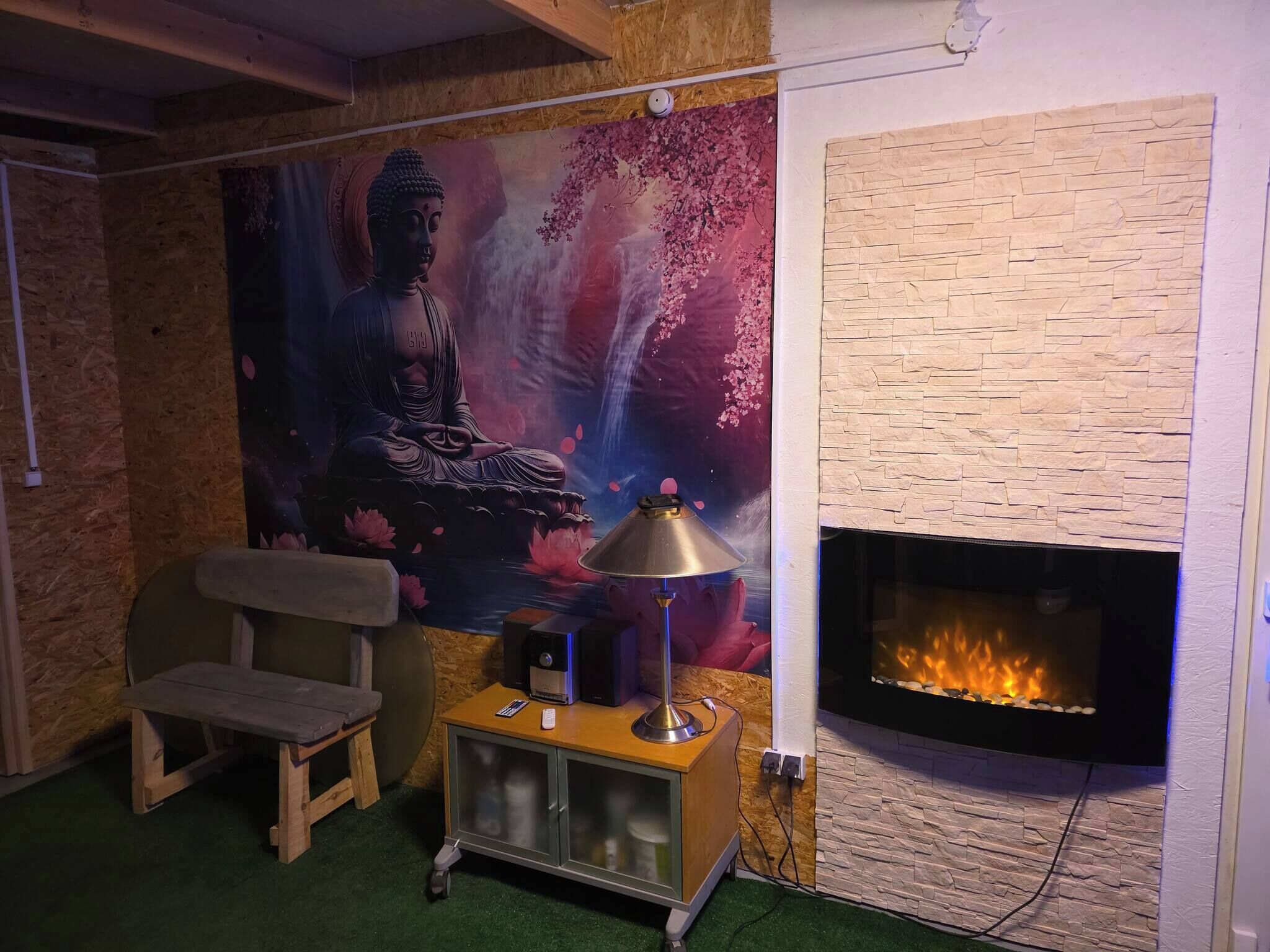 A room with a Buddha mural, a fireplace, a bench, and a small table with a lamp.
