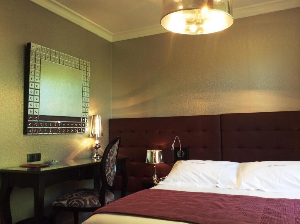 Habitación de hotel con cama, escritorio, silla, mesita de noche, lámpara y una lámpara fija en la pared.
