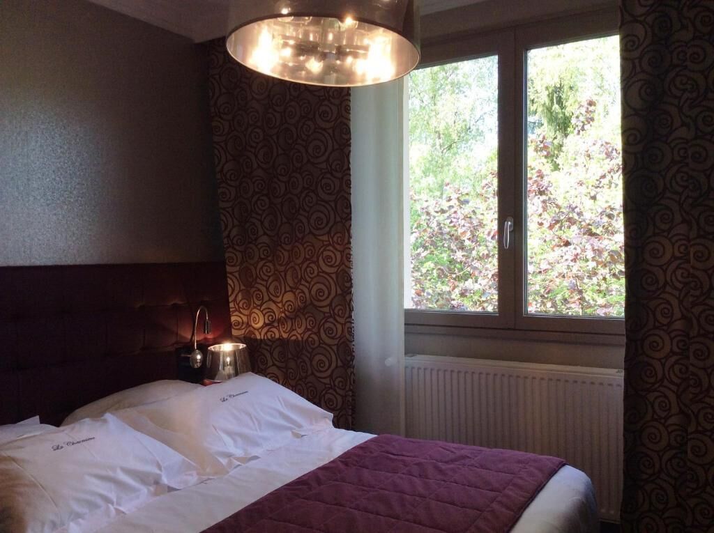 A hotel room with a bed, a bedside table with a lamp, a window with a view of greenery, and patterned curtains.