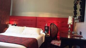 Habitación de hotel con un cabecero tapizado en rojo, una cama con ropa blanca, un escritorio con lámpara y una silla con cojín estampado.