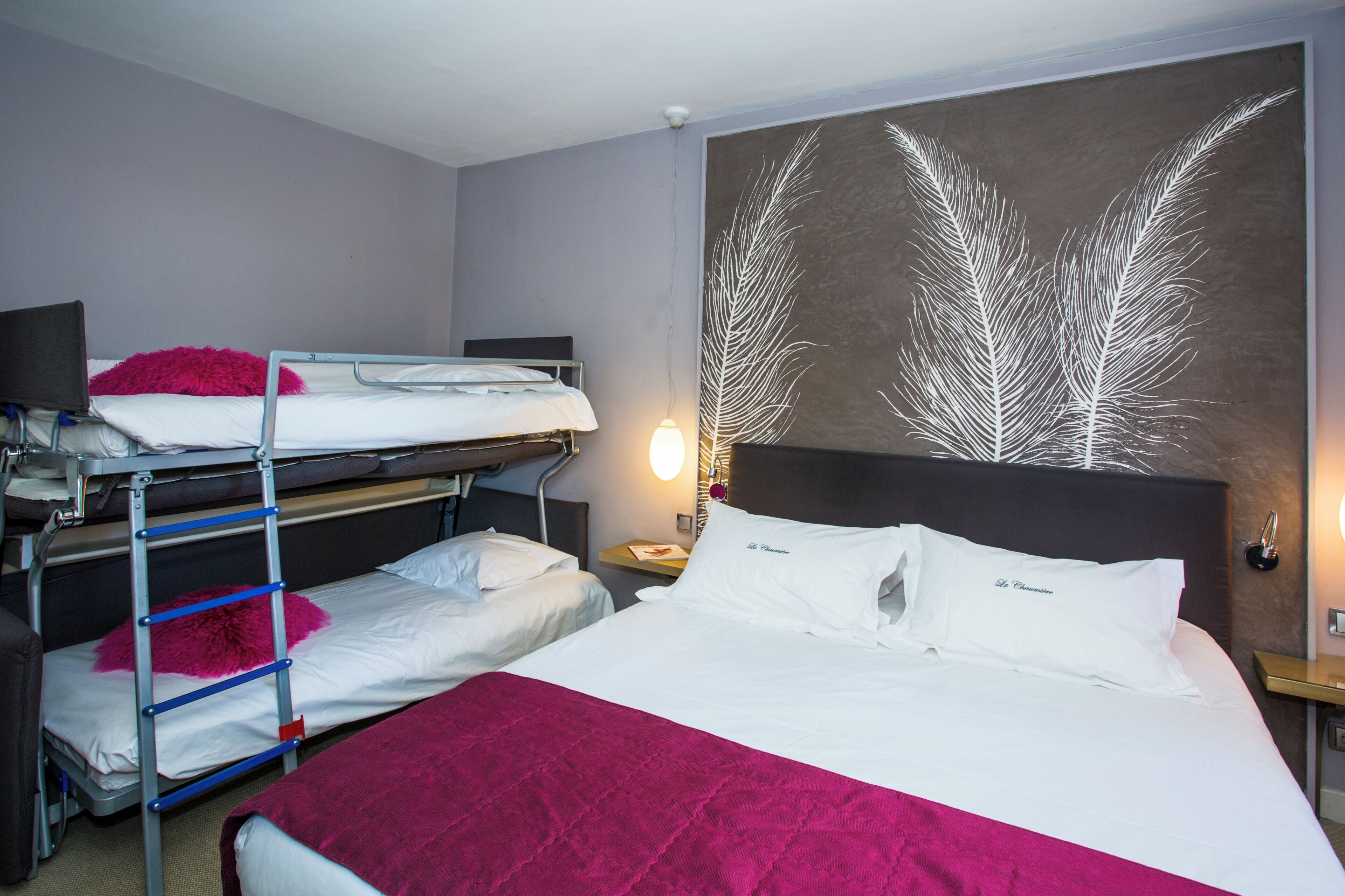 A hotel room with bunk beds, a large bed, a nightstand, a wall-mounted lamp, and a decorative wall piece.