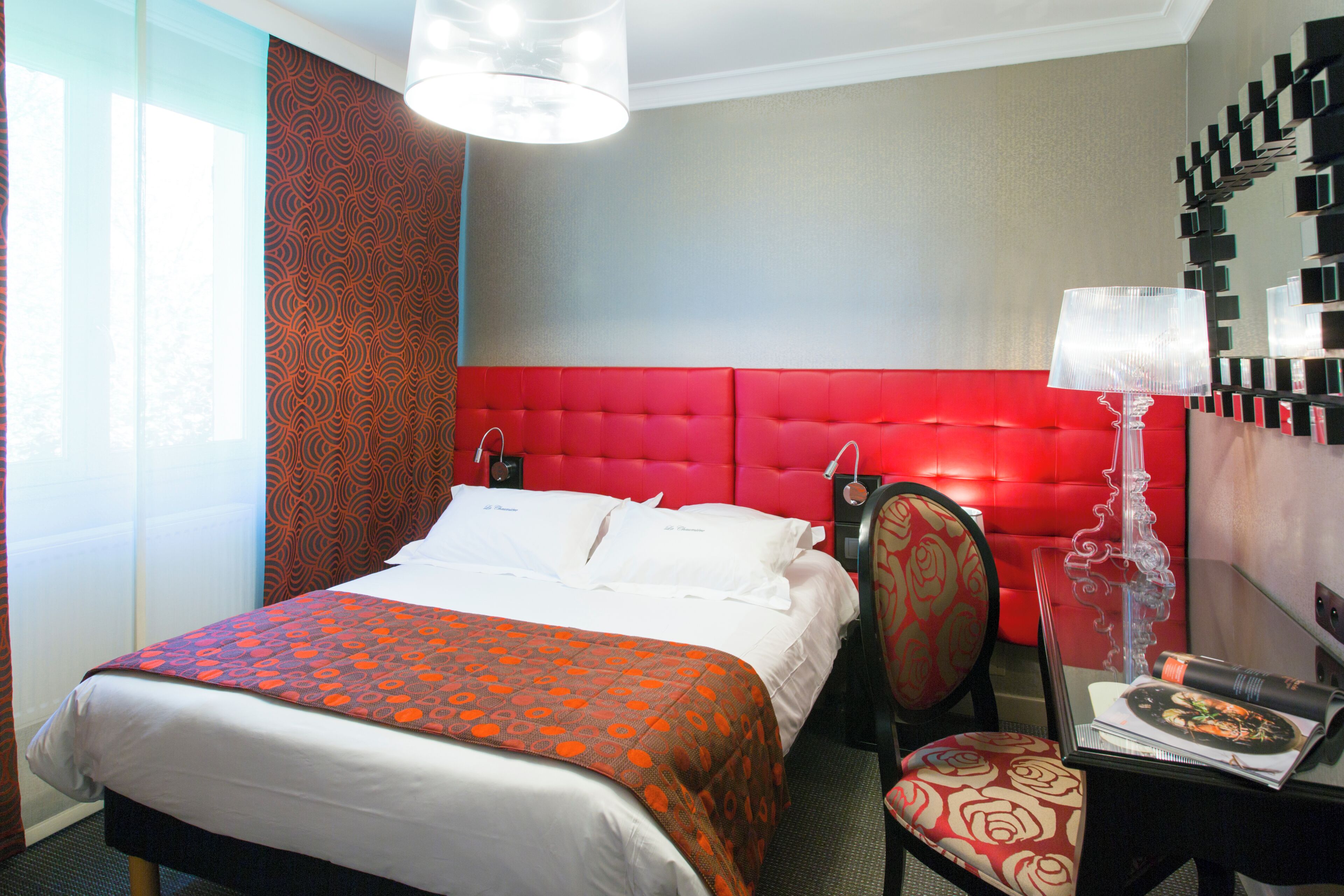A hotel room with a red headboard, a bed with white linens, a red patterned chair, a small table with a lamp, and a wall-mounted shelf with decorative items.
