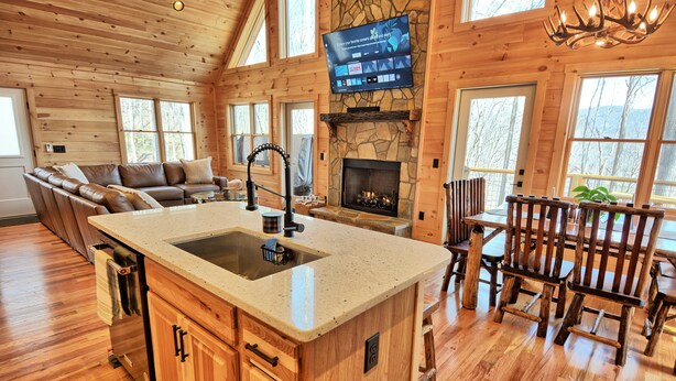 Interior de una cabaña de madera con chimenea de piedra, un televisor montado en la pared y un comedor con muebles de madera.