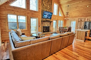 A log cabin interior with a large wooden sofa, a stone fireplace, a dining area, and a kitchen with a refrigerator. - Experience luxury at It'll Do Cabin in Todd, NC—spacious 4BR, mountain views, great amenities! (Todd)