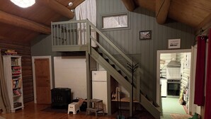 Ein rustikales Blockhaus-Interieur mit Schlafzimmer im Zwischengeschoss, einer Küchenzeile, einem Holzesstisch und einem Bücherregal.